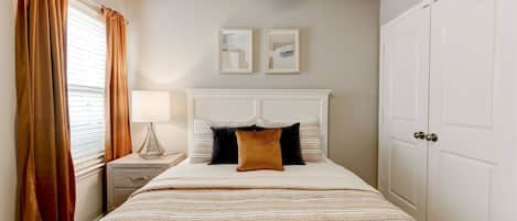 A bedroom with a bed, nightstand, lamp, two framed pictures on the wall, and a wardrobe.
