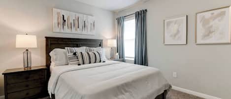 A bedroom with a bed, nightstands, a headboard, artwork, and a window with curtains.