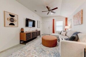 Ein Wohnzimmer mit Fernseher, einer Couch, einem Hocker, einem gemusterten Teppich und einem Deckenventilator.