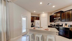 A modern kitchen with dark wood cabinets, a central island with stools, and a white countertop. - Brand New Home in Temple (Temple)