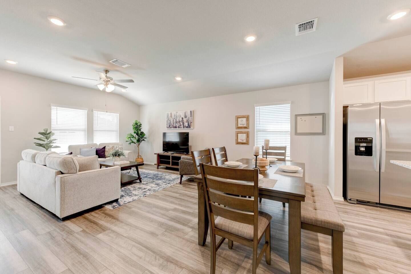 A modern living room with a sofa, coffee table, and TV, a dining area with chairs and a table, and a kitchen with a refrigerator.