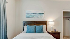 Une chambre d’hôtel avec un lit, une table de chevet, une lampe, un tableau représentant une plage et une armoire.
