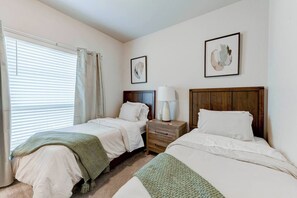 Habitación de hotel con dos camas, una mesita de noche con lámpara, una ventana con cortinas y dos cuadros en la pared.