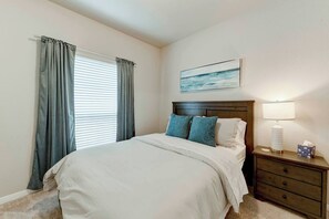 Une chambre à coucher avec un lit, une table de chevet, une lampe, un tableau et une fenêtre avec des rideaux.