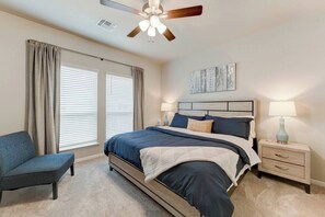 Ein Schlafzimmer mit einem Bett, einem Sessel, einem Nachttisch, einer Lampe, einem Deckenventilator und einem Fenster mit Vorhängen.