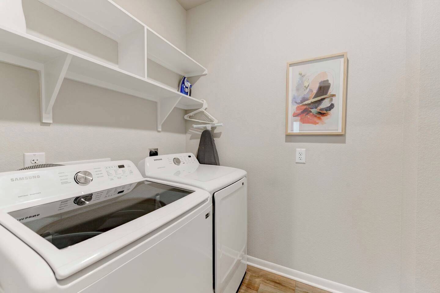 A laundry room with a Samsung washer and dryer, a shelf above the washer, and a framed picture on the wall.
