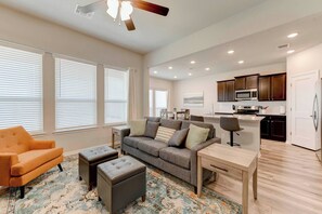 A modern living room with a sofa, ottoman, and armchair, a kitchen with dark wooden cabinets and a refrigerator, and a dining area with a table and chairs.