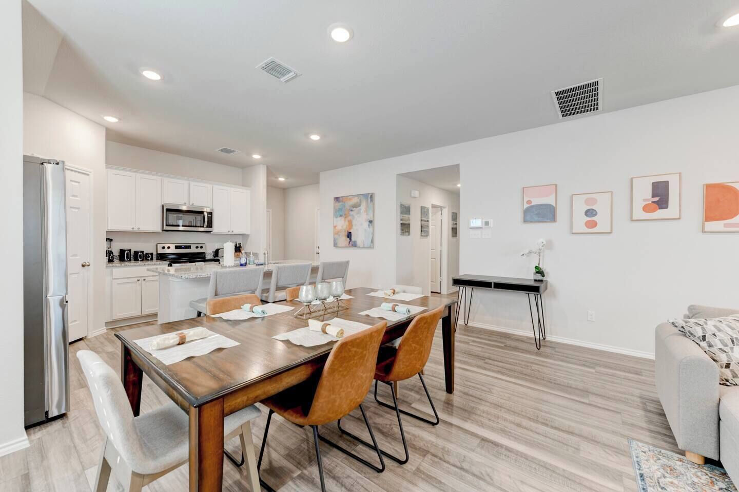 A modern kitchen with white cabinets, a dining area with a wooden table and chairs, and a living area with a sofa and artwork on the wall.
