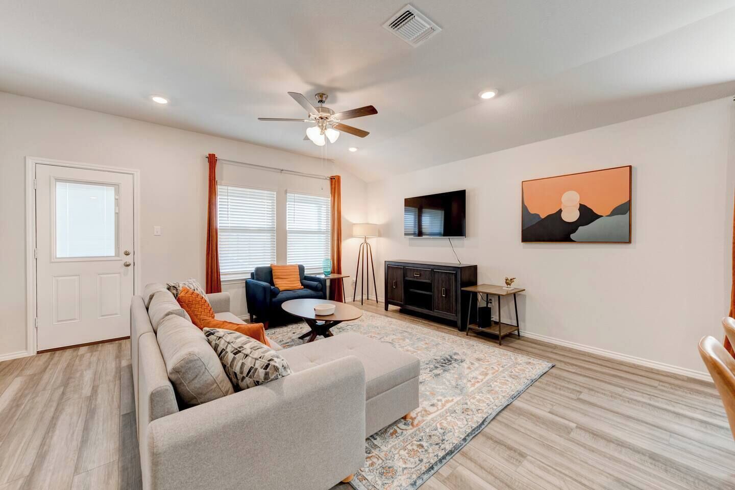 A living room with a sofa, armchair, coffee table, TV, artwork, and a ceiling fan.