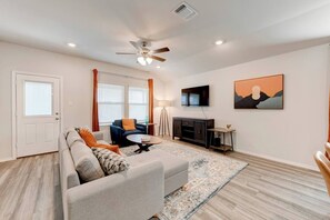A living room with a sofa, armchair, coffee table, TV, artwork, and a ceiling fan.