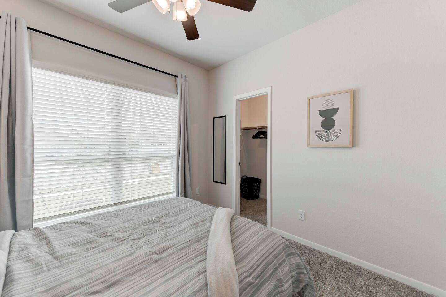A bedroom with a bed, a ceiling fan, a window with blinds, a door leading to a closet, and a framed picture on the wall.