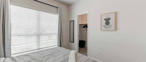 Quarto com cama, ventilador de teto, janela com persianas, porta que dá para um armário e uma moldura com imagem na parede.