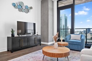 Un salon moderne doté d’un téléviseur à écran plat, de meubles en bois foncé, d’une table basse ronde en bois, d’un canapé bleu et offrant une vue sur la ville grâce à de grandes fenêtres.