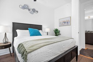 A bedroom with a bed, two bedside tables, a nightstand, a wall-mounted artwork, a potted plant, and a view of the bathroom. - 84 East Avenue, 2303 (Austin)