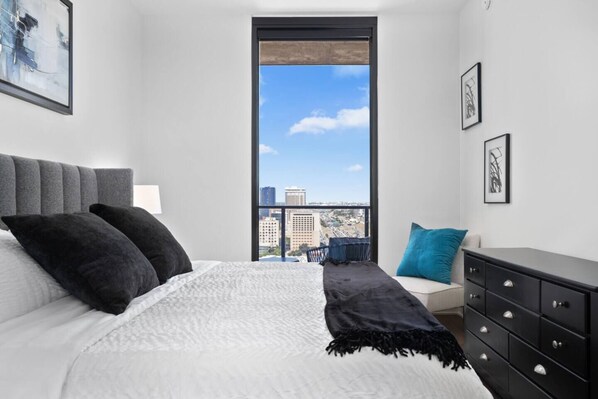 A modern bedroom with a large bed, a view of the city through a window, and a dresser with drawers.