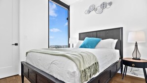Ein modernes Schlafzimmer mit einem großen Bett, einem Nachttisch, einer Lampe und Blick auf den Himmel durch ein Fenster.