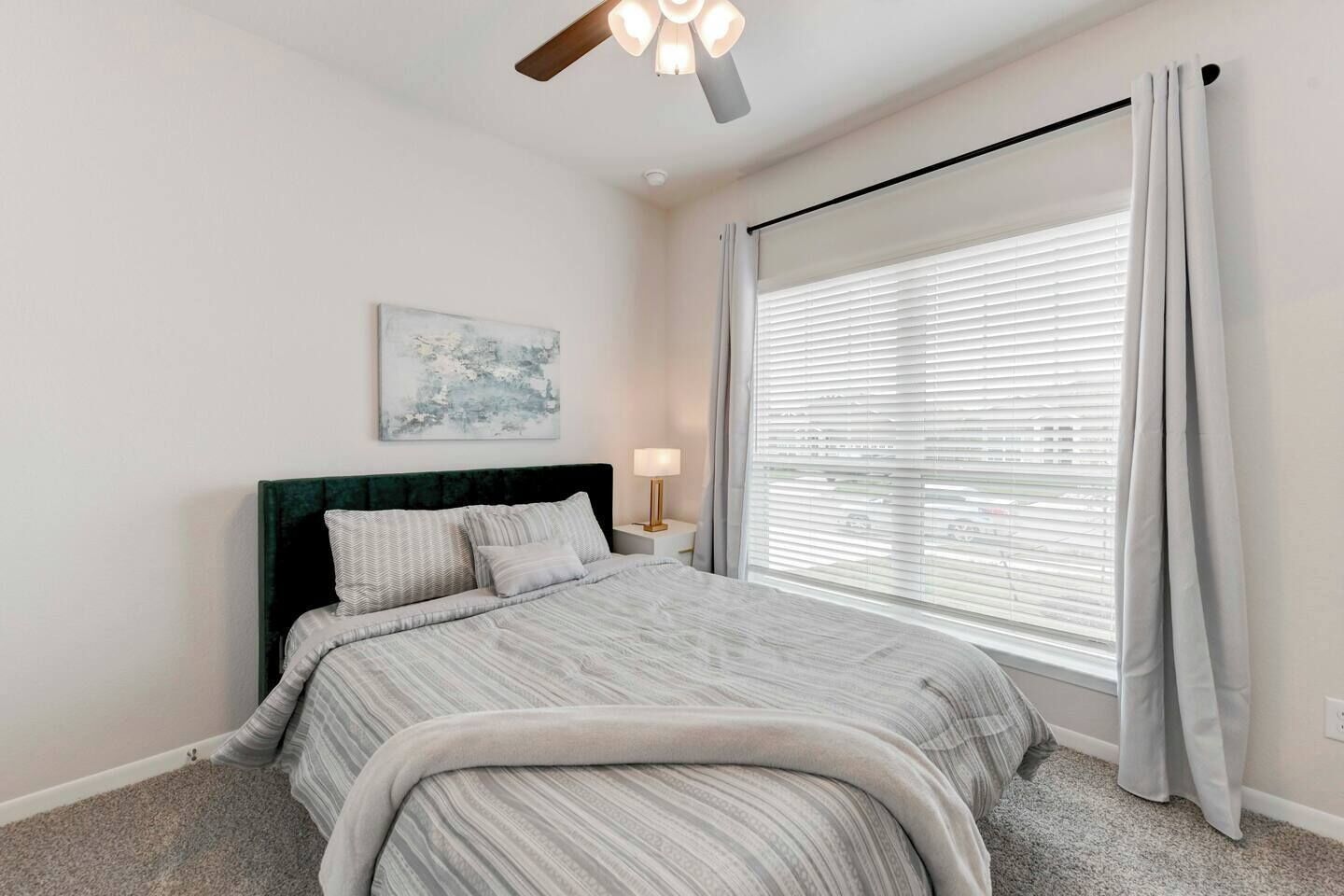 A bedroom with a bed, a nightstand, a lamp, a ceiling fan, and a window with blinds.