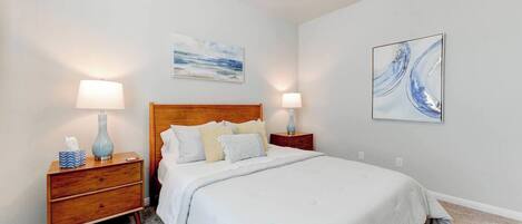 A bedroom with a wooden bed, two bedside tables, a nightstand, a wall-mounted painting, and a carpeted floor.