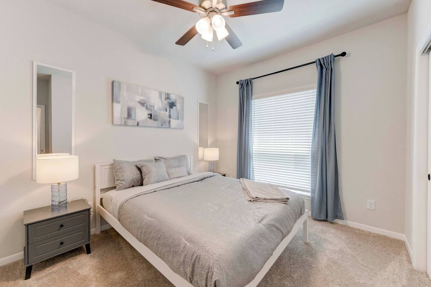 Une chambre à coucher comprenant un lit, des tables de chevet, un ventilateur de plafond, une fenêtre avec des rideaux et un miroir fixé au mur.