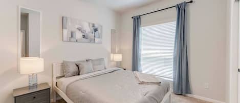 Une chambre à coucher comprenant un lit, des tables de chevet, un ventilateur de plafond, une fenêtre avec des rideaux et un miroir fixé au mur.