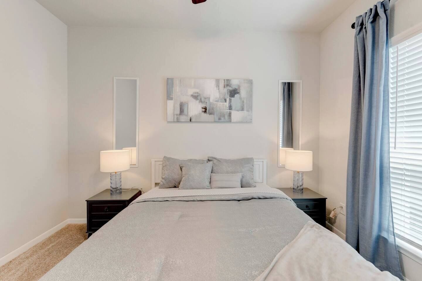 A bedroom with a bed, two bedside tables, a mirror, and a window with curtains.