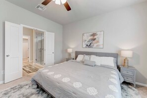 A bedroom with a large bed, two bedside tables with lamps, a door leading to a bathroom, and a ceiling fan.