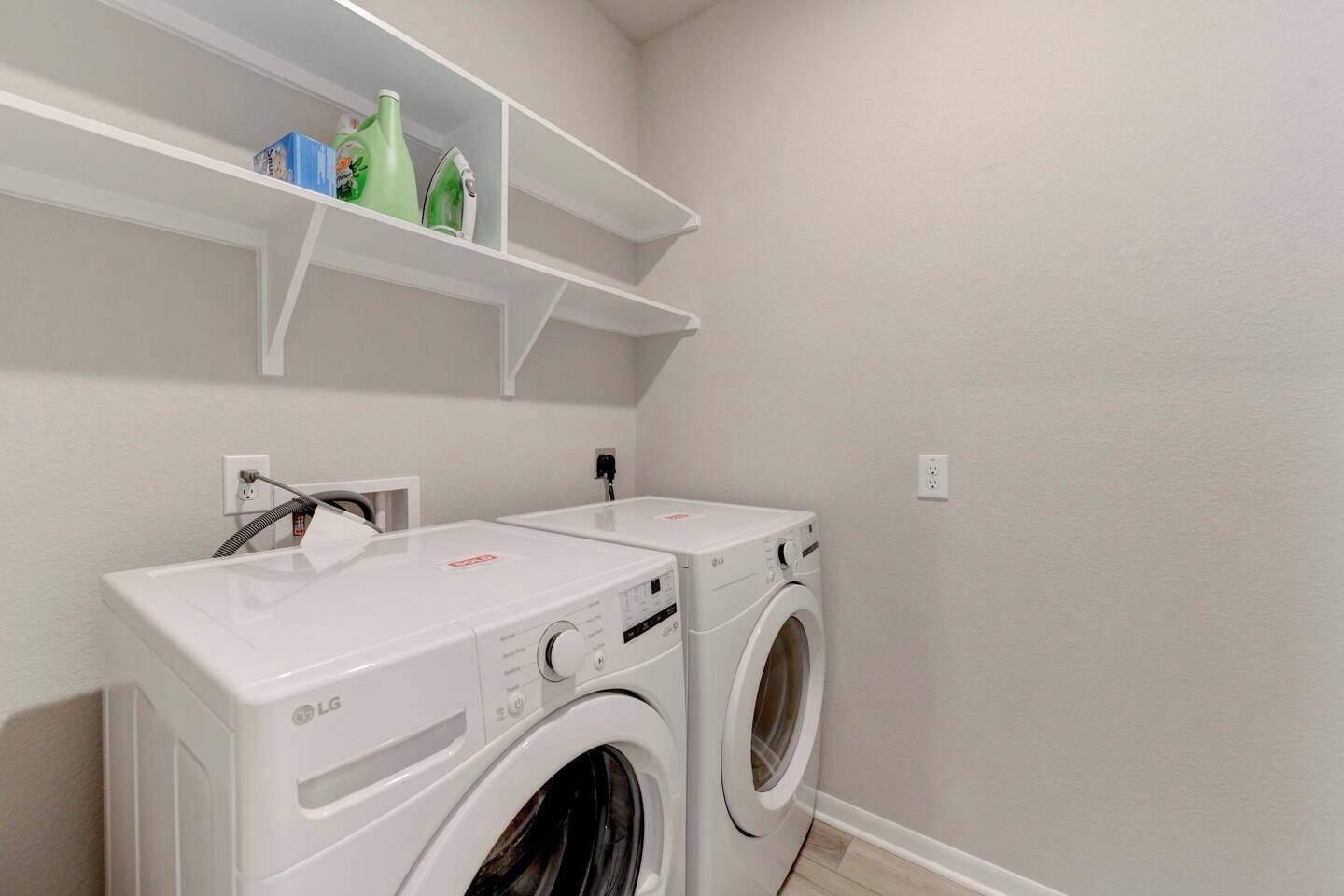 A laundry room with two LG washing machines, a shelf above with cleaning supplies, and a power outlet on the wall.