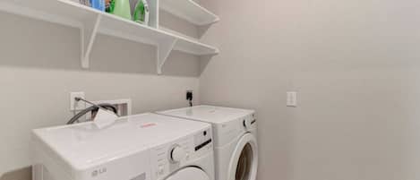 A laundry room with two LG washing machines, a shelf above with cleaning supplies, and a power outlet on the wall.