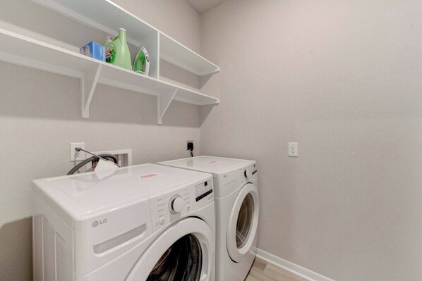 Un cuarto de lavandería con dos lavadoras LG, un estante superior con productos de limpieza y un enchufe en la pared.
