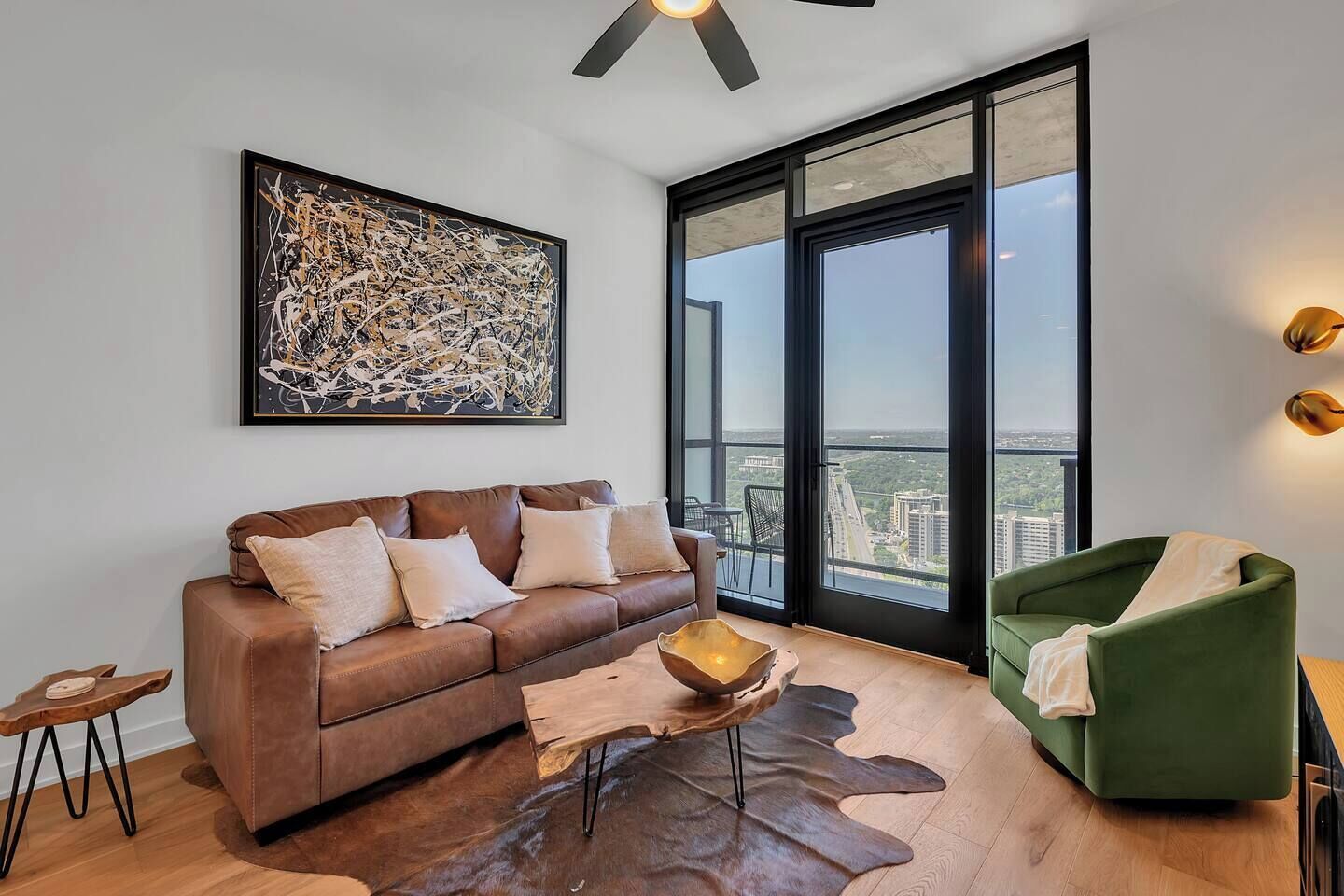 A modern living room with a leather sofa, a green armchair, a large abstract painting, and a city view through floor-to-ceiling windows.