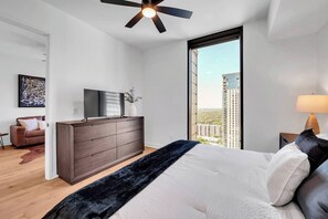 A modern bedroom with a large bed, a dresser, a TV, and a city view.