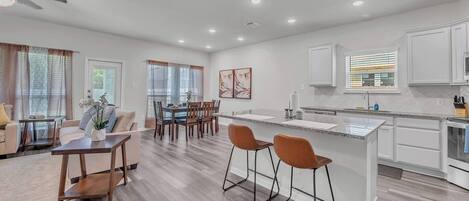 A modern kitchen with an island, dining area with chairs, and a living room with a sofa and coffee table.