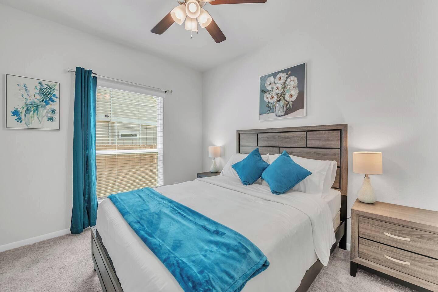 A bedroom with a bed, a ceiling fan, a nightstand, a lamp, a window with curtains, and two wall art pieces.