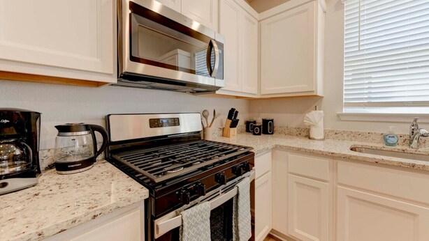 A modern kitchen with white cabinetry, a stainless steel range hood, a coffee maker, and a sink with a window above it.