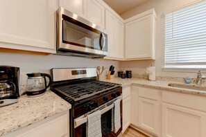 A modern kitchen with white cabinetry, a stainless steel range hood, a coffee maker, and a sink with a window above it. - Round Round Temporary Housing (Round Rock)