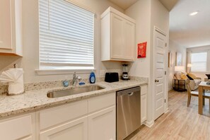 A kitchen with white cabinets, a granite countertop, a dishwasher, a blender, and a window with blinds. - Round Round Temporary Housing (Round Rock)