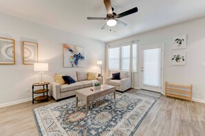 A living room with a beige sofa, a coffee table, a patterned rug, and a ceiling fan. - Round Round Temporary Housing (Round Rock)