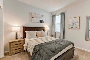 A bedroom with a bed, two bedside tables, a nightstand, a headboard, a wall-mounted artwork, and a window with curtains. - Round Rock Executive Suite (Round Rock)