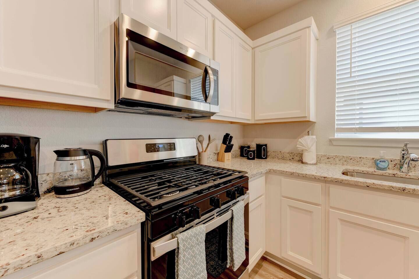 A kitchen with white cabinets, a stainless steel range, a coffee maker, and a window with blinds.