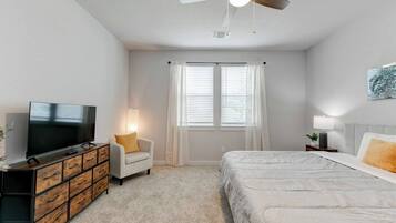 A bedroom with a bed, a TV, a chair, a dresser, a ceiling fan, and a window with curtains.