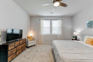 A bedroom with a bed, a TV, a chair, a dresser, a ceiling fan, and a window with curtains.