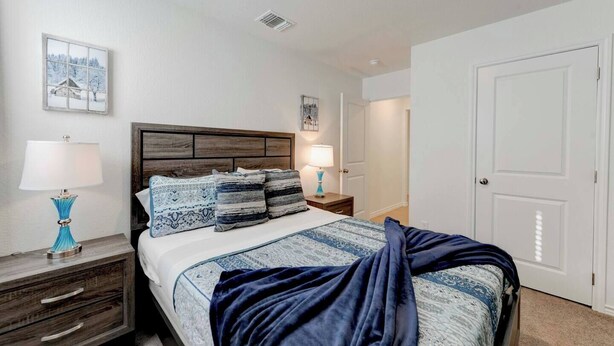 A bedroom with a wooden headboard, a bed with patterned bedding, two bedside tables with lamps, a nightstand, and a door.