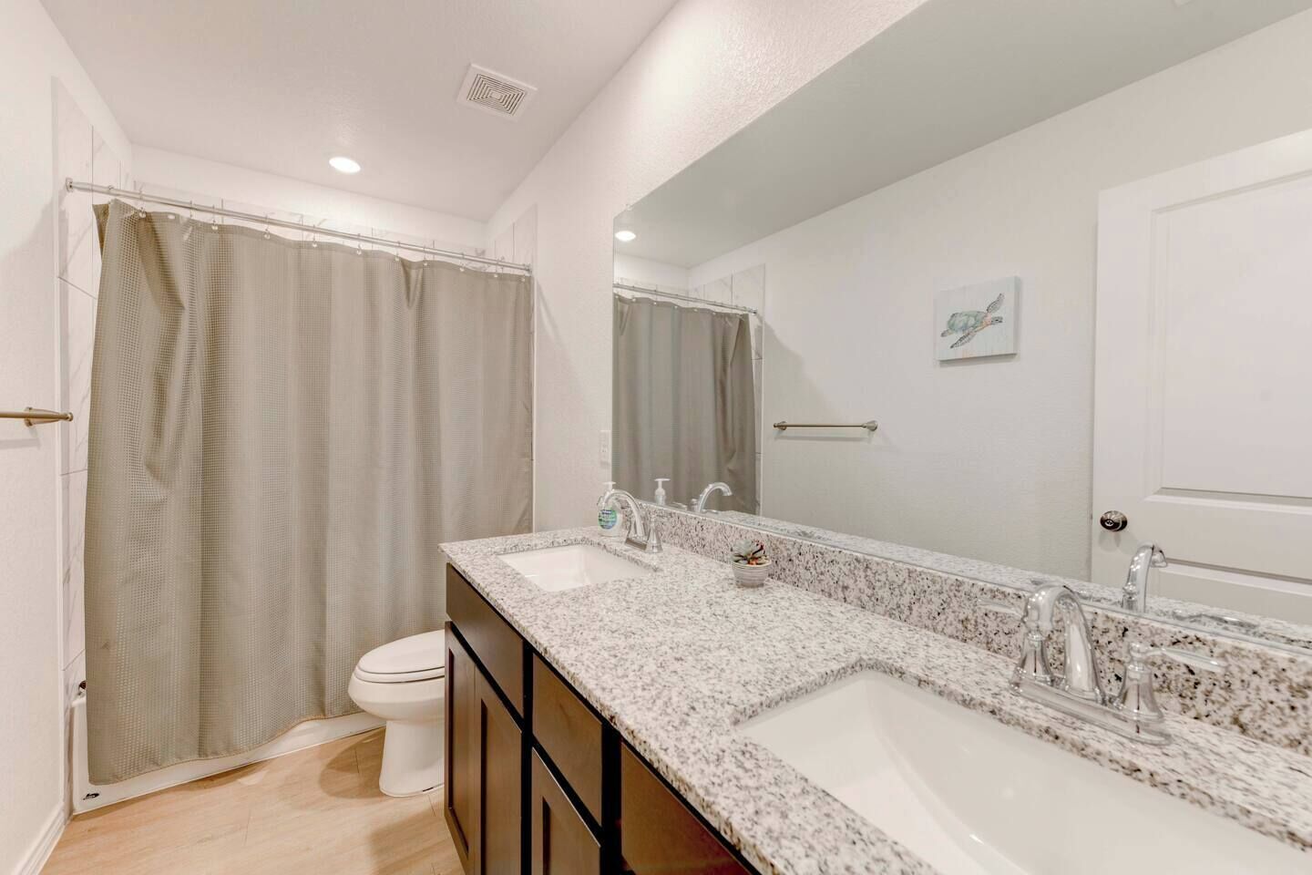 A bathroom with a double sink, a toilet, a shower curtain, a large mirror, and a towel rack.