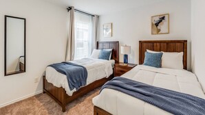 A hotel room with two beds, a mirror, a window with curtains, a bedside table with a lamp, and a picture on the wall.
