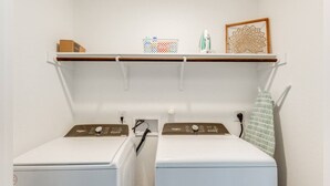 A laundry area with two front-loading washers and dryers, a shelf above with a framed picture, and a power outlet on the wall. - New Braunfels Serene Gateway (New Braunfels)