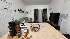 A modern living room with a sofa, coffee table, and a TV. There is a fireplace, a plant, and a window with curtains.