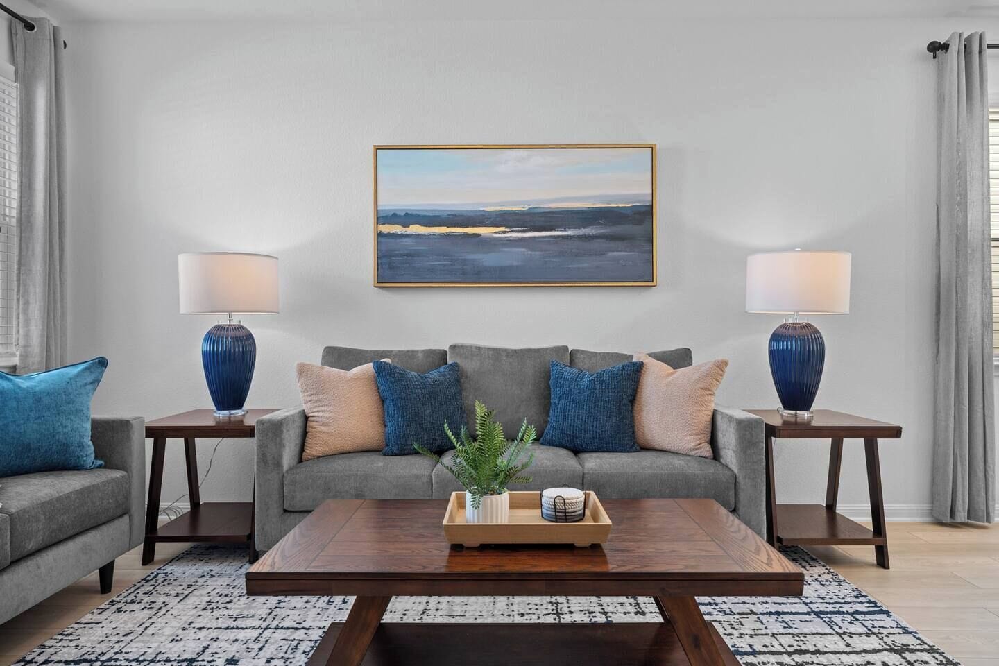Un salon avec un canapé gris, deux tables d’appoint avec des lampes, une table basse et un tableau représentant un paysage, encadré, accroché au mur.