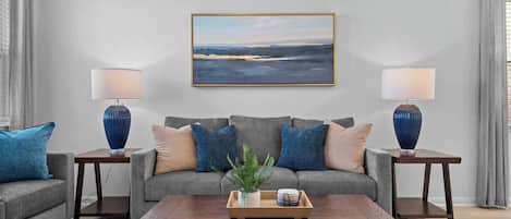 A living room with a grey sofa, two side tables with lamps, a coffee table, and a framed landscape painting on the wall.