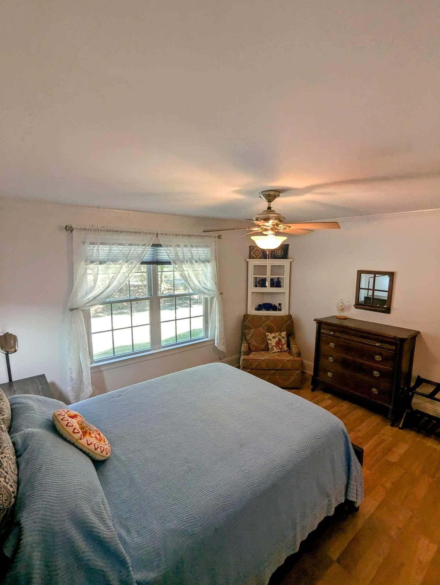 Une chambre à coucher comprenant un lit, une chaise, une commode, un ventilateur de plafond et une fenêtre avec des rideaux.