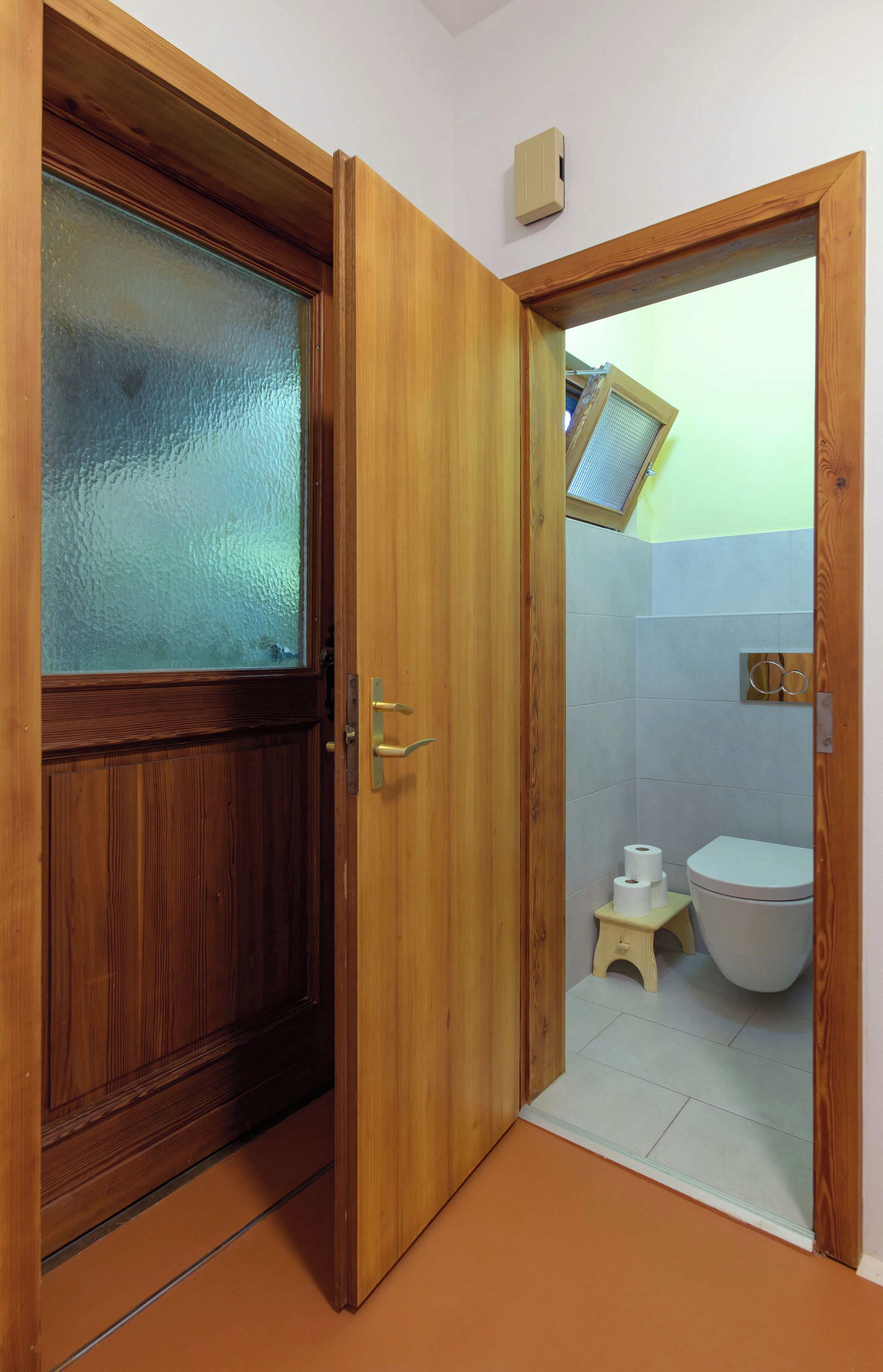 Puerta de madera que da a un baño con un inodoro, un taburete pequeño y una ventana.